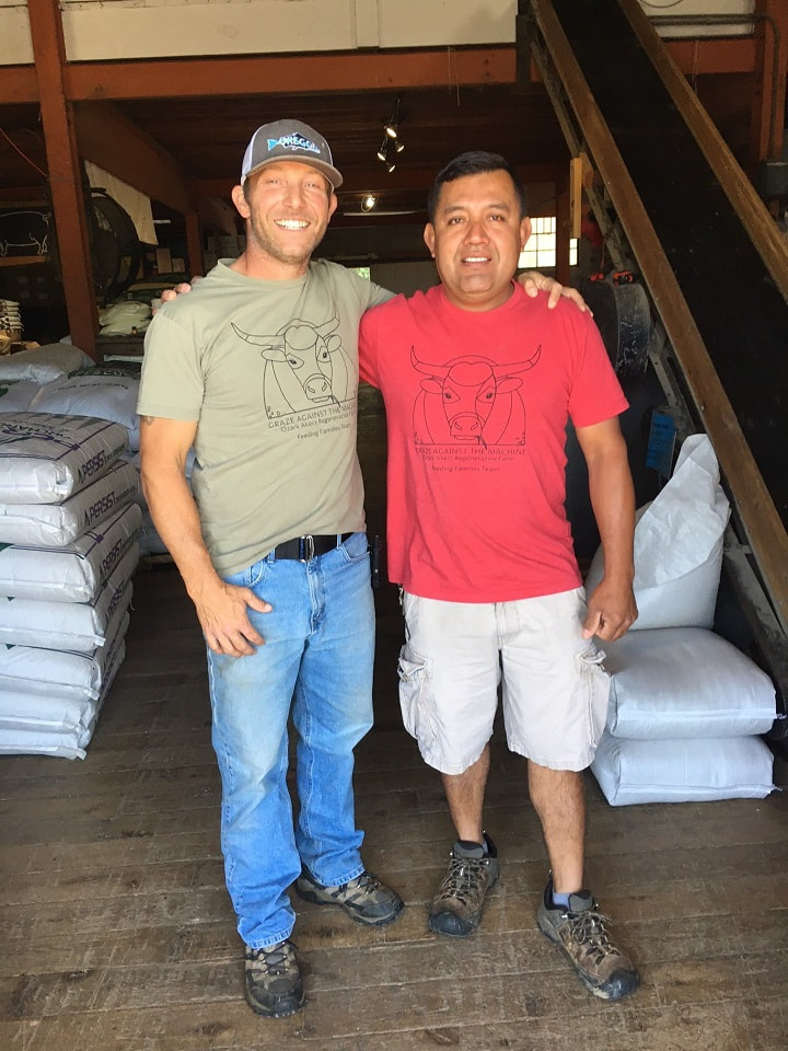 Josh and Antonio standing infont of sacks of organic feed at Country Farm and Home farm supply, they are both wearing a Graze Against The Machine, Feeding Families t-shirt