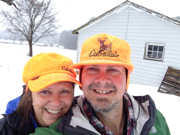 Sue and Mike wearing orange Cabelos hats stand in front of the shed during a snow storm